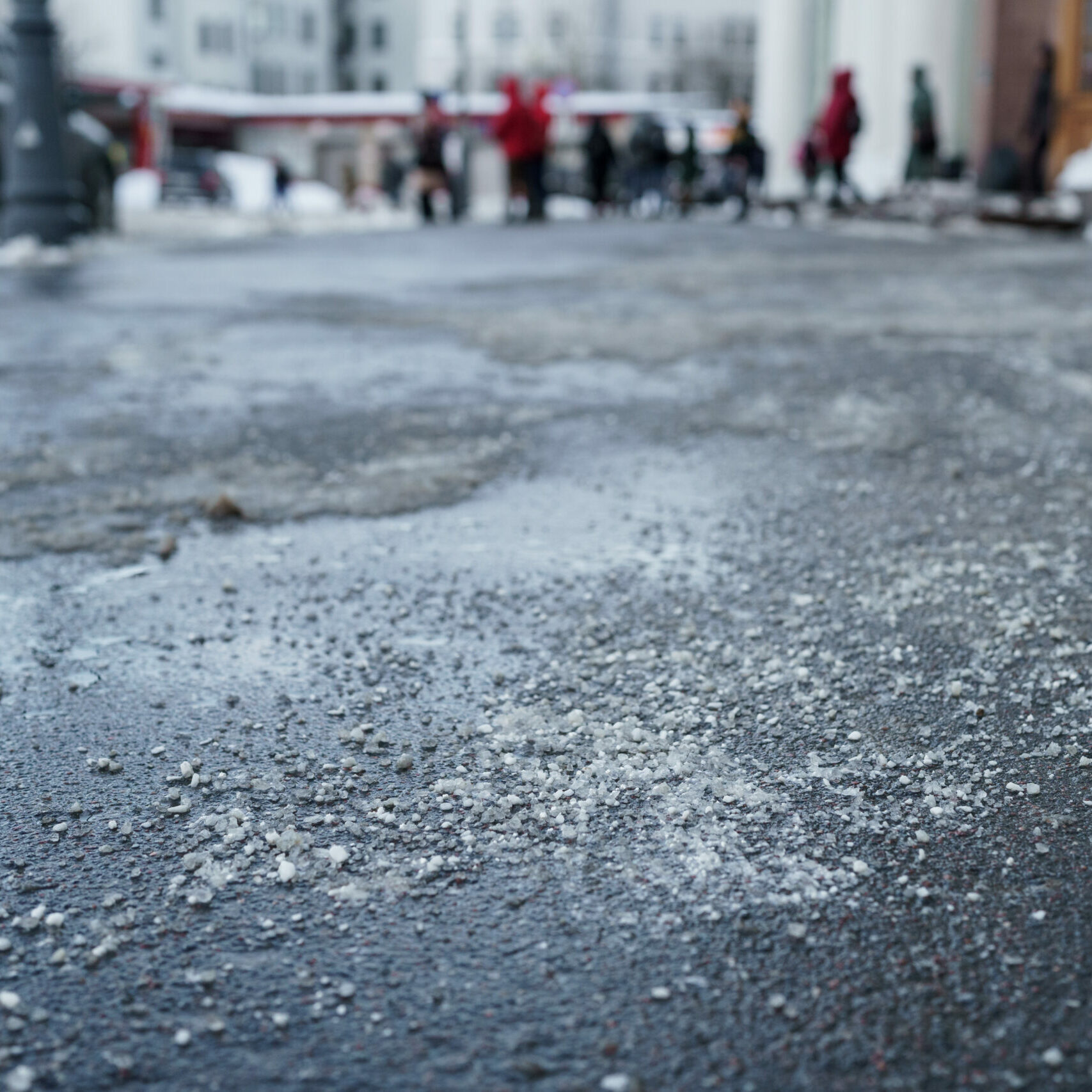 De-icing chemical reagent on the road will make the road safe in winter time. Pavement is sprinkled with technical salt, focus on foreground, people on blurred background.